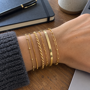Gold six-chain bracelet stack on an adult wrist beside a notebook · AI-generated illustrative demo image