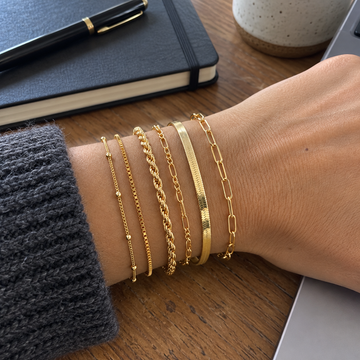 Gold six-chain bracelet stack on an adult wrist beside a notebook · AI-generated illustrative demo image