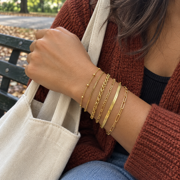 Gold six-chain bracelet stack on an adult wrist holding a canvas tote · AI-generated illustrative demo image